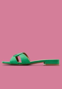 Sandale en suède vert au design plat, avec deux straps croisés et une pointe ouverte, reposant sur une semelle fine sur un fond rose.