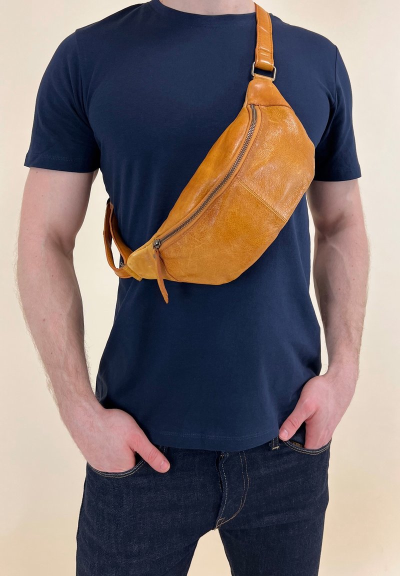 Homme portant un t-shirt bleu marine et un jean avec un sac bandoulière en cuir beige porté sur la poitrine, mains dans les poches.