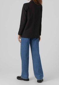 Person med lockigt hår i svart kavaj, blå vida jeans och svarta skor som står med ryggen mot kameran på en grå bakgrund.