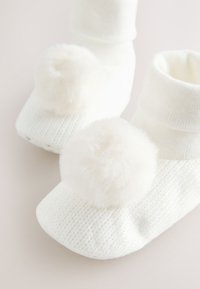 Chaussons blancs en tricot avec un pompon doux et duveteux à l'avant. Dotés d'un revers côtelé et d'une semelle texturée pour une meilleure adhérence.
