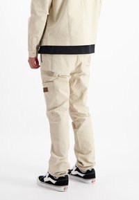 Beige canvas broek met een recht-toe-recht-aan ontwerp, voorzien van zijzakken en een bruine leren accent. Gecombineerd met zwart-witte sneakers.
