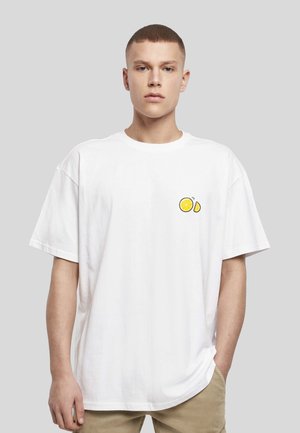 Νέος άντρας με κοντά μαλλιά που φορεί μια oversized άσπρη μπλούζα με μικρό σχέδιο λεμονιού στο αριστερό στήθος και μπεζ παντελόνι.