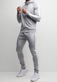 Tuta grigio chiaro in tessuto elasticizzato, con felpa con cappuccio con zip e pantaloni affusolati con strisce laterali bianche e dettagli del marchio.