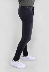 Jeans de denim negro con un corte ajustado y un ligero estiramiento. Detalle notable de bolsillo con cremallera en el lado derecho. Llevados con zapatillas blancas.