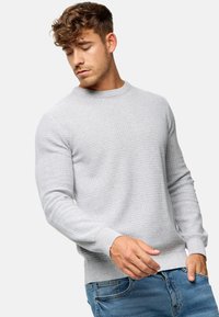 Pull en maille gris clair avec un col rond et des poignets côtelés, affichant un motif texturé sur l'ensemble. Associé à un jean bleu.