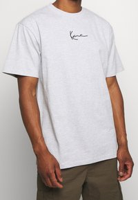 Camiseta gris de manga corta hecha de una mezcla de algodón. Presenta un logo en cursiva negra en el lado izquierdo del pecho. Diseño simple y de corte relajado.