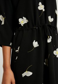 Vêtement noir à motifs floraux blancs avec touches jaunes. Coupe décontractée, taille élastique avec lien ajustable, manches trois-quarts.