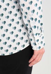 Long-sleeve shirt in white fabric featuring a repeating heart pattern in teal and light blue. Standard cuff with a button closure.