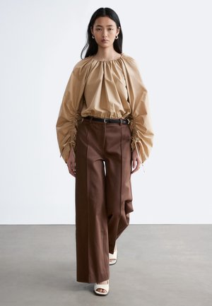 Žena v béžovej blúzke s nazbieranými dlhými rukávmi, hnedých širokých nohaviciach s čiernym opaskom a bielych sandáloch s otvorenou špičkou, kráčajúca vpred.