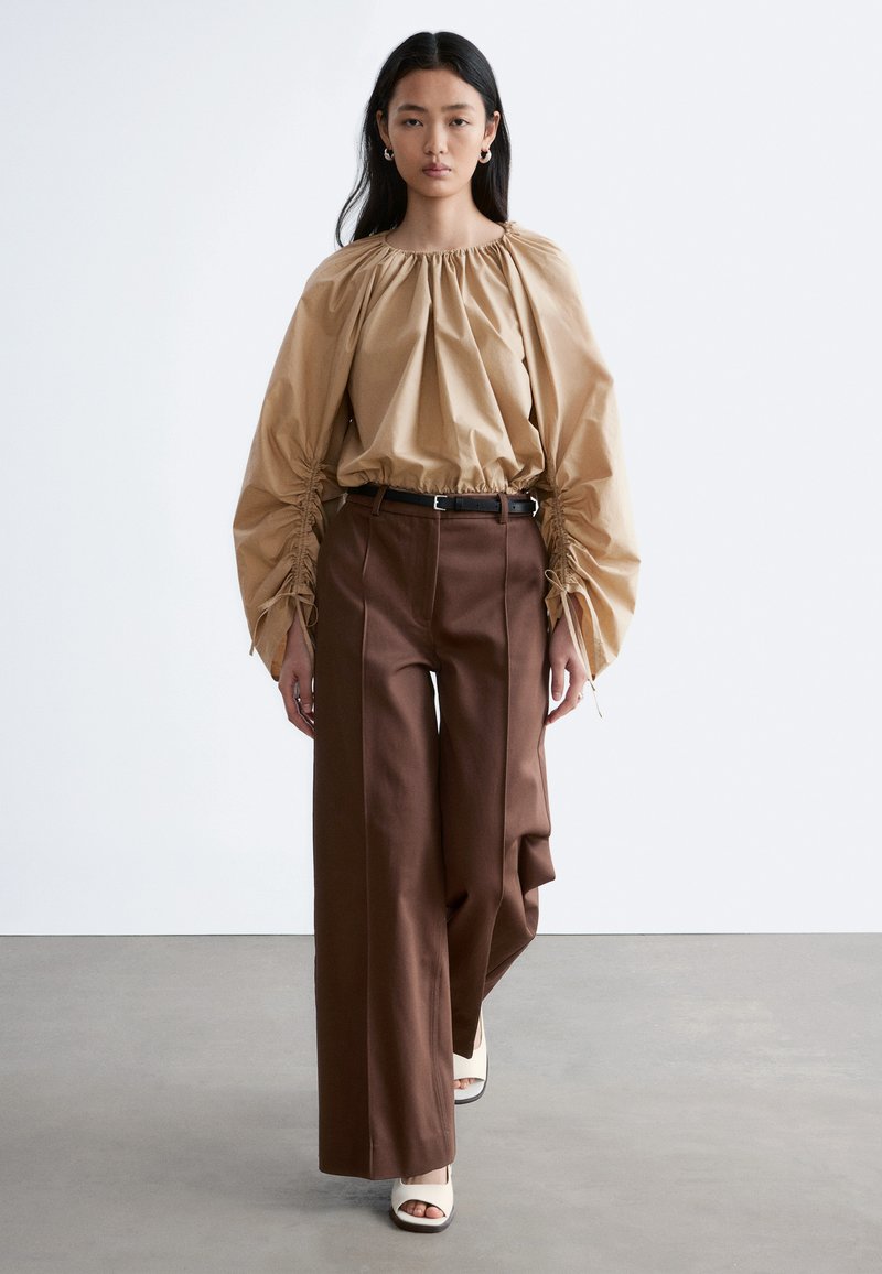 Femme portant un chemisier beige à manches longues froncées, un pantalon large marron avec une ceinture noire, et des escarpins blancs à bout ouvert, marchant vers l'avant.