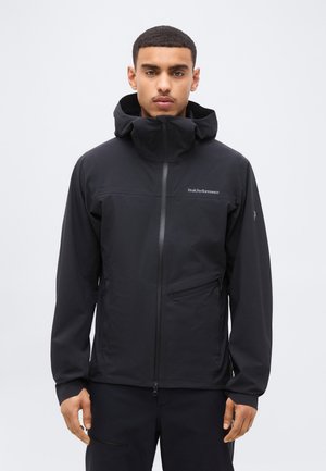 Muž oblečený v čiernej kapucňovej bunde Peak Performance s predným zipsom a vreckami na zips, stojaci pred jednoduchým svetlým pozadím.