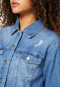 Mujer con cabello rizado vistiendo una chaqueta de mezclilla azul con botones de metal y un pequeño rasguño desgastado cerca del bolsillo del pecho.