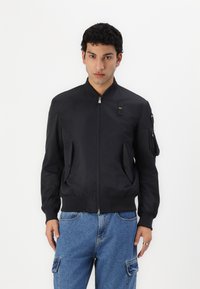 Giubbotto bomber nero con zip frontale, polsini a costine, due tasche anteriori e toppa sulle spalle. Il materiale appare liscio e leggero.