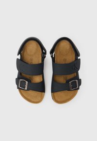 Μαύρα σανδάλια με δύο ρυθμιζόμενους ιμάντες, μεταλλικό αγκράφα και μαλακό καφέ πέλμα. Τ ο ανώτερο υλικό φαίνεται λείο και συνθετικό.