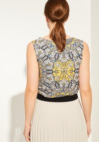 Ærmeløs bluse med et flerfarvet mønstret stof med gule, blå og sorte accenter og et sort taljebånd. Tekstureret rygdesign.