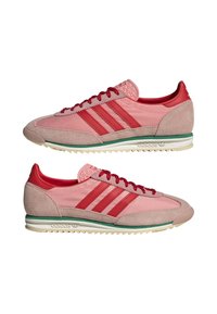 Adidas cipők, amelyek világos rózsaszín és piros anyagok keverékével készültek, három piros csíkkal, velúr részletekkel és zöld szegélyezésű fehér talppal.