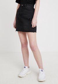 Osoba oblečená v černé distressed mini sukni z denimu s nepravidelným lemem a bílými teniskami, stojící na jednobarevném světlém pozadí.