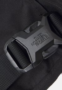Černý nylonový pásek s odolnou černou plastovou sponou, na které je vyraženo logo "The North Face". Texturovaná tkanina viditelná vedle spony.