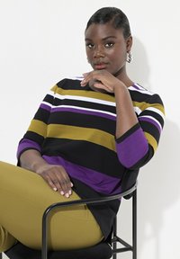 Randig topp med svart, vitt, lila och olivgrönt; rund halslinje; 3/4 ärmar. Modellen sitter med olivgröna byxor.