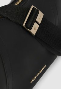 Bolso de cuero negro con una correa texturizada negra y herrajes dorados. Cuenta con una cremallera visible y un detalle de logotipo en relieve.