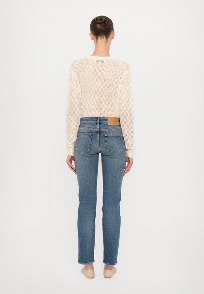 Leichter, cremfarbener, offen gestrickter Pullover mit langen Ärmeln, kombiniert mit blauen, schmal geschnittenen Jeans, die an der hinteren Tasche einen braunen Lederpatch haben.