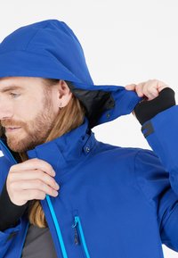 Uomo con lunghi capelli e barba che regola il cappuccio di una giacca impermeabile blu con una cerniera blu chiaro su uno sfondo bianco.