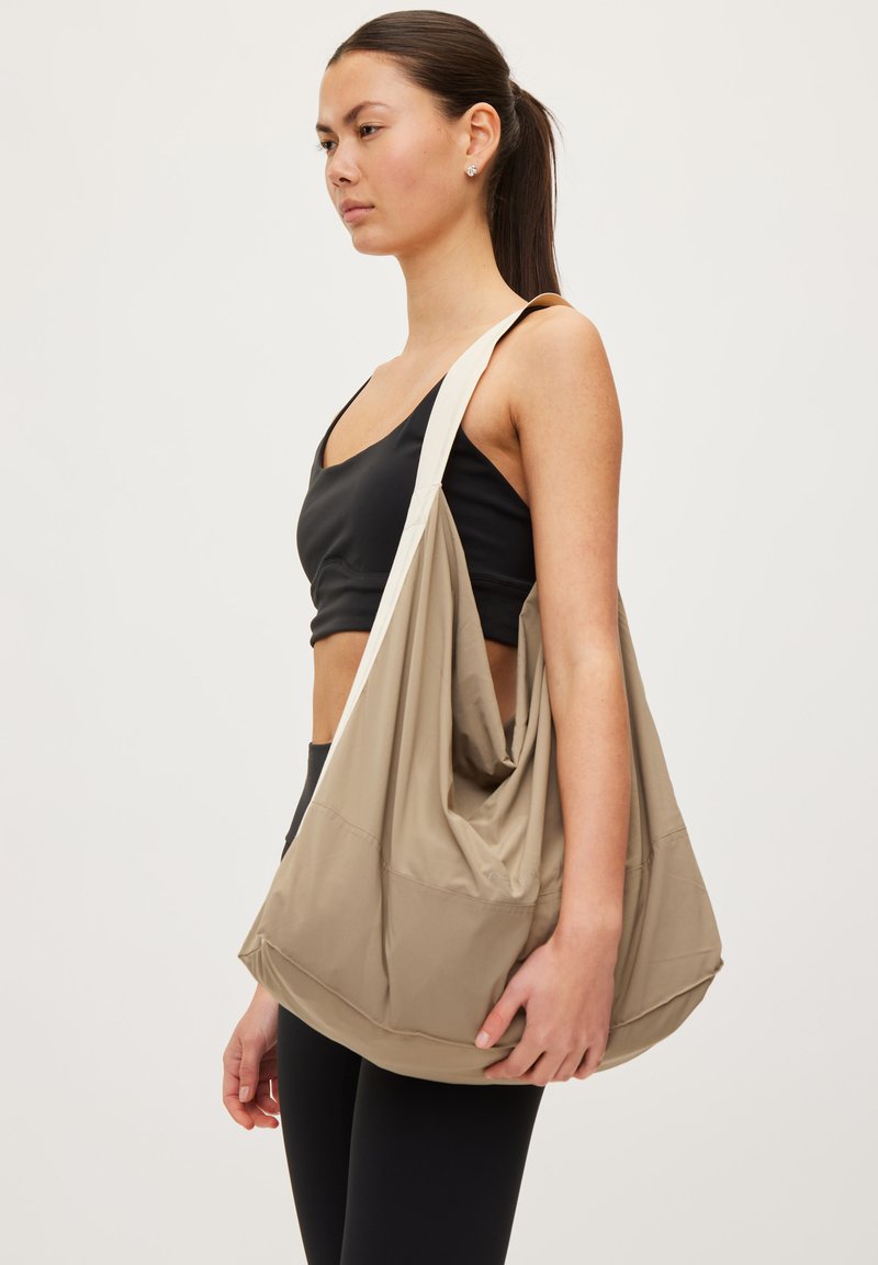 Kobieta ubrana w czarne sportowe ubrania trzyma dużą beżową, luźną torbę na ramię na jednolitym jasnym tle.