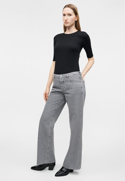 Femme debout, mains dans les poches, portant un haut noir ajusté, un jean évasé taille haute gris et des bottines à talons noires sur un fond uni.