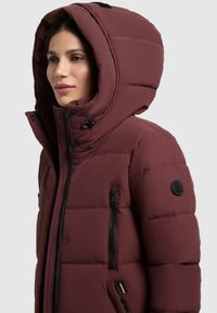 Chaqueta acolchada color burdeos con una gran capucha, bolsillos con cremallera y un parche redondo de logo negro. Presenta una textura acolchada y un grueso aislamiento.