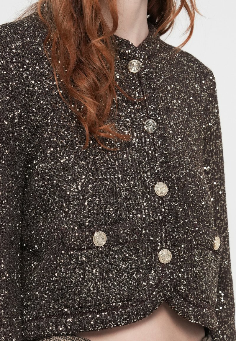 Schwarze, mit Pailletten besetzte, kurze Jacke mit silbernen Blumenknöpfen und Klappentaschen, getragen von einer Person mit welligem roten Haar.