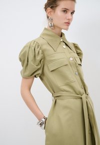 Abito camicia verde oliva con maniche a sbuffo, chiusura frontale con bottoni, due tasche sul petto e vita cinturata. Un bracciale argento al polso completa il design.