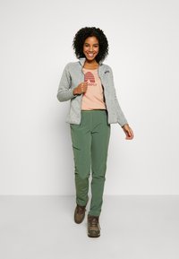 Grau Fleecejacke über einem hellrosa Shirt mit Grafik, kombiniert mit grünen Hosen und braunen Wanderstiefeln. Modell lächelt und geht nach vorne.