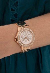 Roségoldene Uhr mit einem mit Edelsteinen besetzten Gehäuse, weißem Zifferblatt und drei Unterzifferblättern. Das Metallglied-Armband sorgt für Langlebigkeit; glatte Textur und polierte Oberfläche.