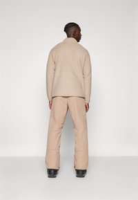 Veste en polaire beige avec un col, assortie à un pantalon beige clair. Le pantalon dispose d'une poche latérale et d'un détail de logo. Chaussures noires visibles.