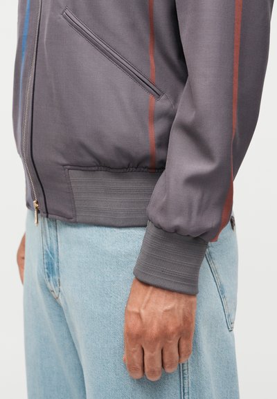 Πλευρική όψη ενός ατόμου που φορά γκρι ζακέτα με φερμουάρ και κόκκινες ρίγες και ανοιχτό μπλε τζιν, δείχνοντας τις λεπτομέρειες της μανσέτας και της τσέπης του μπουφάν.