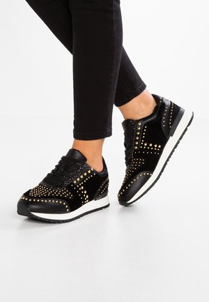 Füße in schwarzen Turnschuhen mit goldenen Nieten, schwarzen Schnürsenkeln und weißen Sohlen, kombiniert mit schwarzen, eng anliegenden Hosen vor weißem Hintergrund.