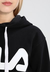 Zwarte hoodie met een voorzak in kangoeroestijl, witte trekkoorden en een opvallend wit grafisch ontwerp op de borst. Zacht katoenen materiaal, comfortabele pasvorm.