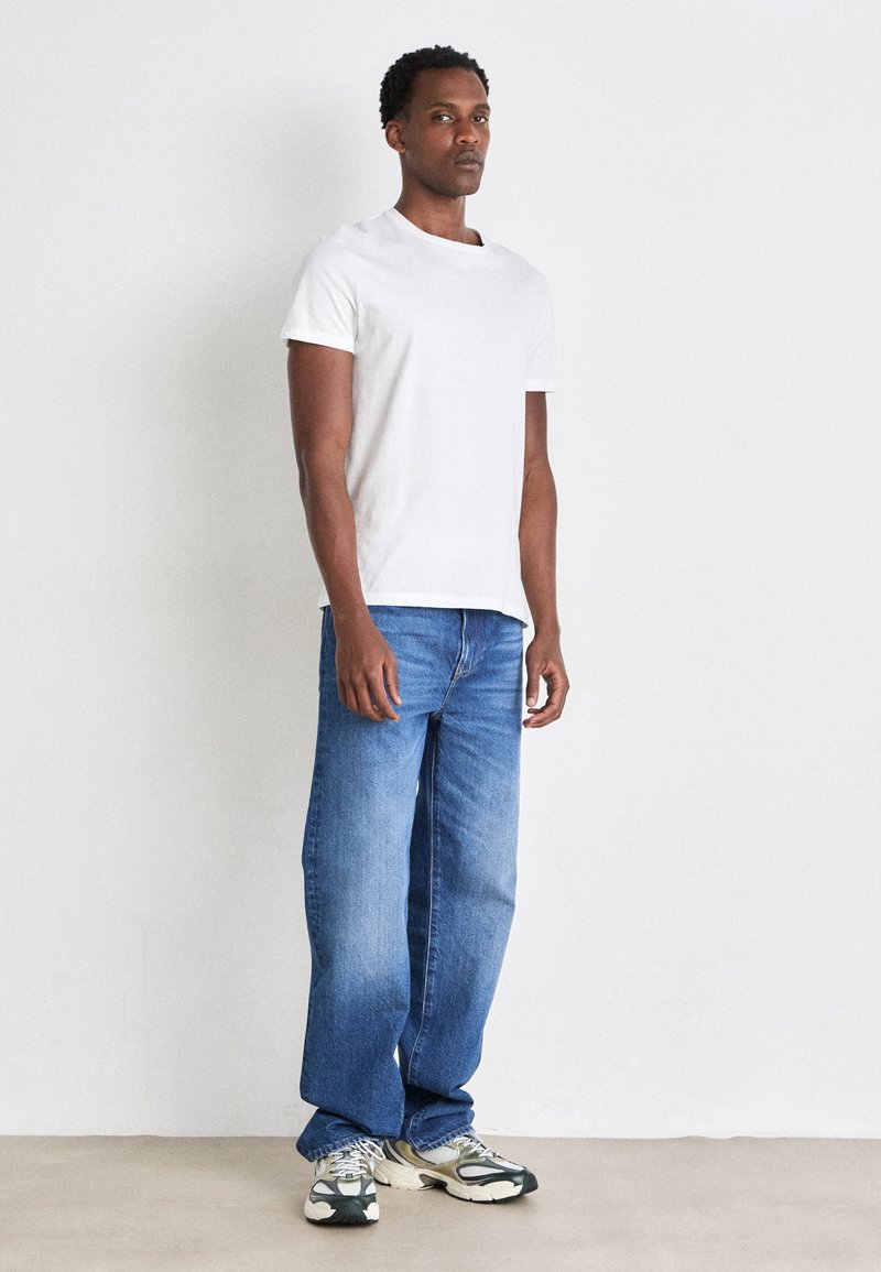 T-shirt bianca in cotone, jeans blu a vestibilità libera, posizionati su uno sfondo chiaro, completati da sneakers chunky multicolori.