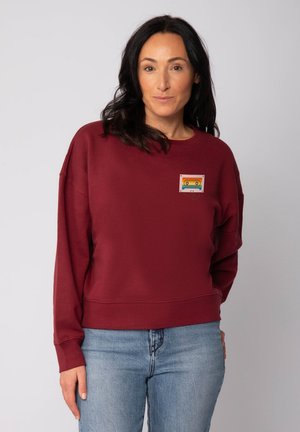 Vrouw met donker haar in een maroon sweatshirt met een kleurrijke cassettebandje grafiek en lichtblauwe spijkerbroek, staande tegen een effen achtergrond.
