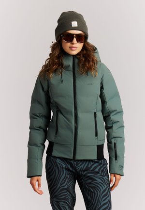 Vrouw met een groene winterjas met kap, een donkergroene muts, grote zonnebril en zwarte broek met donkerblauwe zebrastripes.