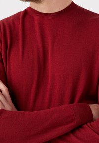 Czerwony sweter z dzianiny z prążkowanym okrągłym dekoltem i długimi rękawami, wykonany z miękkiego materiału o gładkiej fakturze.