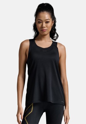 Mujer con cabello largo y oscuro en una coleta alta, vestida con una camiseta deportiva negra sin mangas y leggings negros con detalles dorados, de pie contra un fondo blanco.