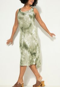Grün gefärbtes, ärmelloses Kleid mit lockerer Passform, seitlichem Schlitz und weichem Stoff. Getragen mit braunen Plattform-Clogs.