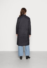 Abrigo largo negro acolchado de corte recto, con cuello alto, combinado con unos vaqueros de pierna ancha azul claro y botas negras cortas.