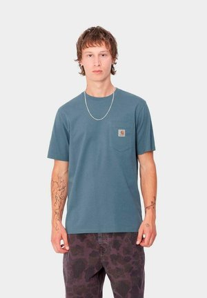 Camiseta de algodón en color verde azulado, de manga corta, con cuello redondo y un bolsillo en el pecho izquierdo. Presenta una etiqueta de logo en el bolsillo y se lleva con pantalones oscuros de estampado.