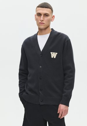 Jonge man met kort haar die een zwart gebreid vest met knopen draagt, met een witte "W" patch over een wit overhemd, staand met handen in de zakken.