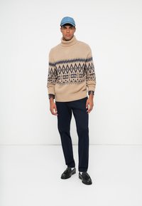Pull à col roulé beige en tricot avec des motifs géométriques, pantalon bleu marine et chaussures noires. L'individu porte une casquette bleu clair.