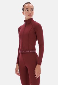 Garš piedurkņu sporta krekls tumši sarkanas krāsas, izgatavots no elastīga auduma. Aprīkots ar augstu apkakli un rāvējslēdzēju priekšpusē. Jostasvietā ir logo lenta kā detaļa.