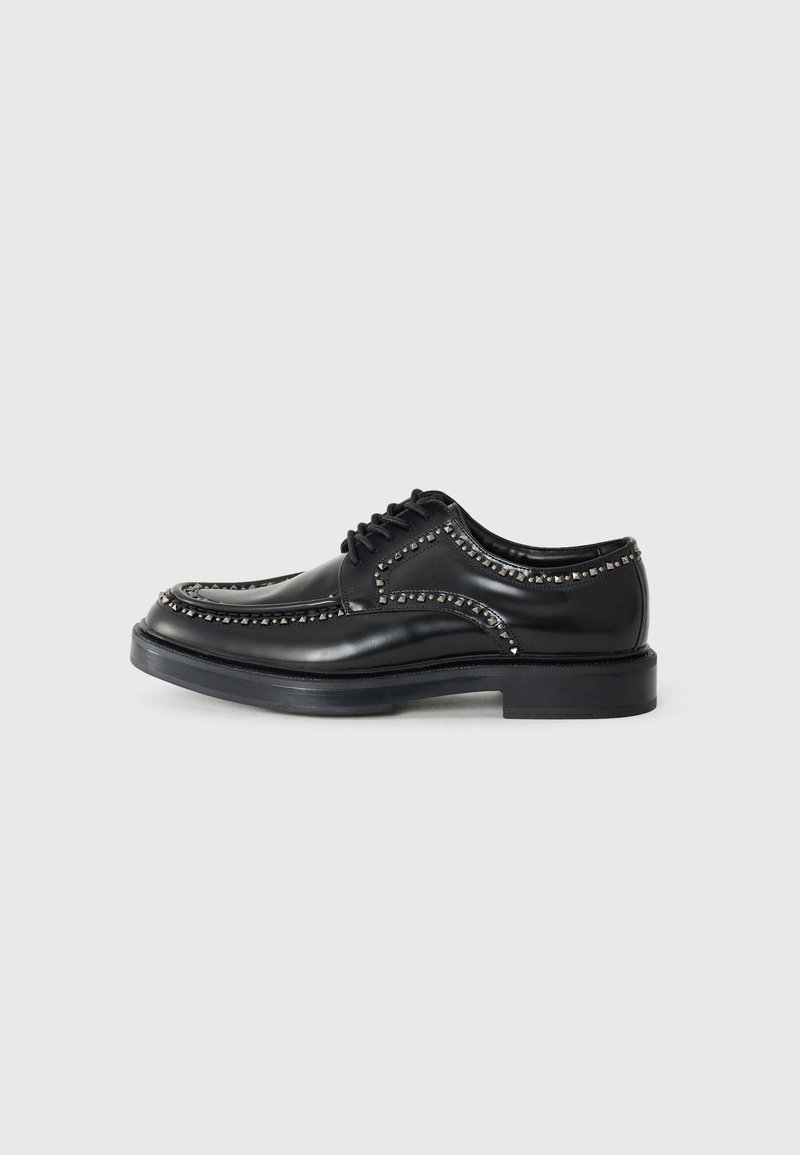 Black leather lace-up shoes with decorative studs along the seams, rounded toe, and a thick, flat black rubber sole.