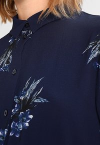 Blusa blu navy con collo a polo e apertura con bottoni, caratterizzata da una stampa floreale in tonalità di blu e nero. Tessuto dalla consistenza liscia.